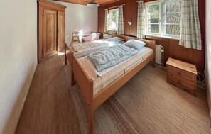 A wooden bed with two pillows and a blanket, a wooden nightstand, a radiator, and a window with curtains.