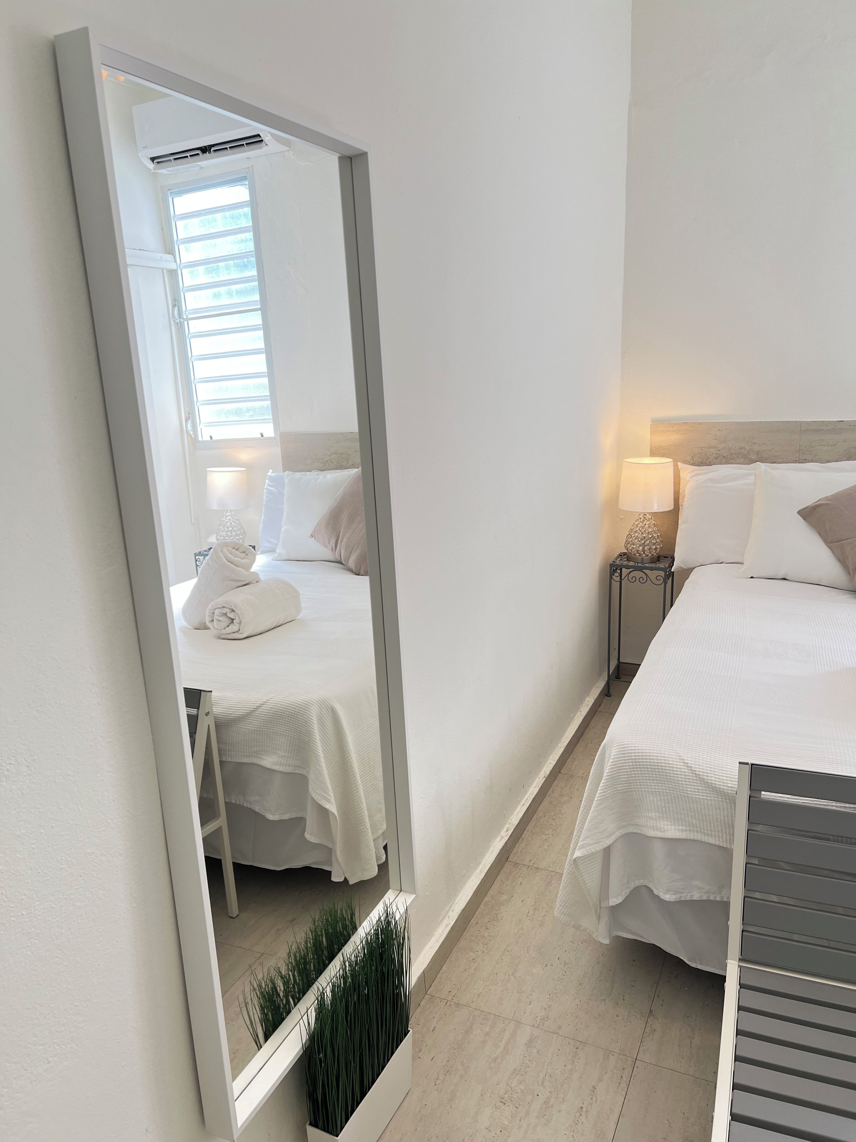 A hotel room with two beds, a large mirror, a bedside table with a lamp, and a window with blinds.