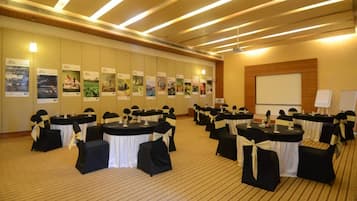 Un salón de conferencias con mesas redondas, sillas negras con cintas blancas, una pantalla de proyector y una pared con carteles.