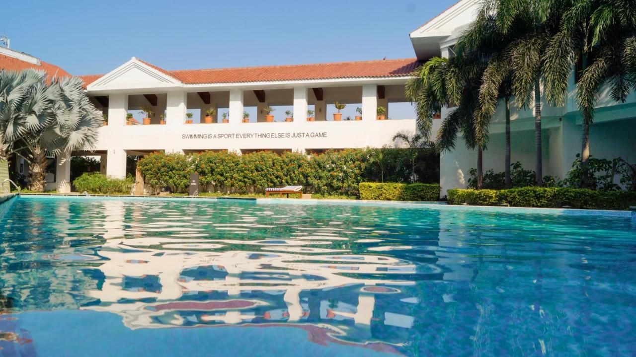 A swimming pool with a clear blue water surface, surrounded by lush greenery and a building with a red-tiled roof.