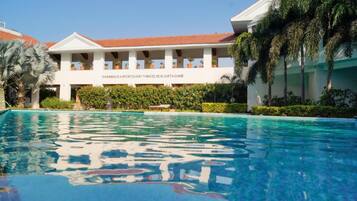 A swimming pool with a clear blue water surface, surrounded by lush greenery and a building with a red-tiled roof.