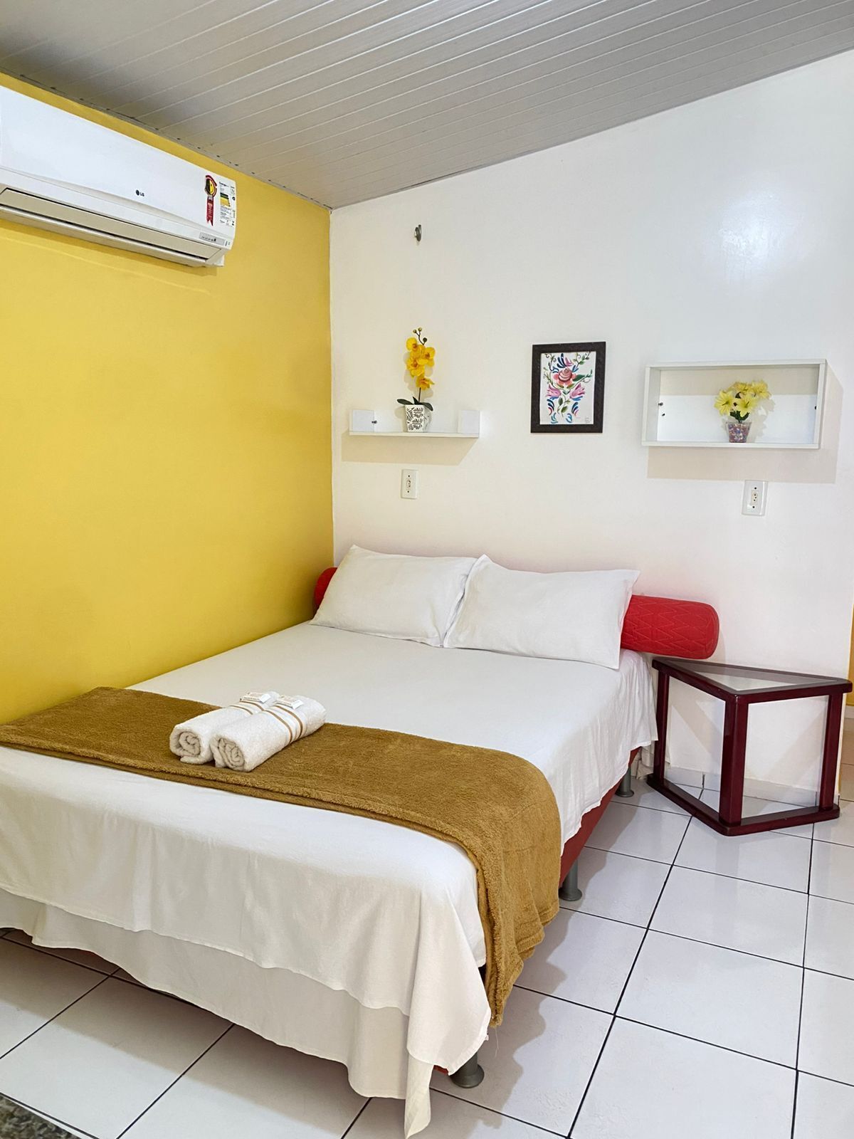 A hotel room with a bed, a bedside table, a wall-mounted air conditioner, and two wall-mounted shelves with decorative items.
