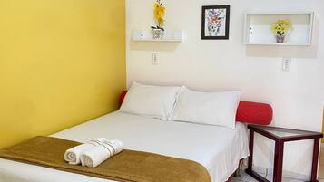 A hotel room with a bed, a bedside table, a wall-mounted air conditioner, and two wall-mounted shelves with decorative items.