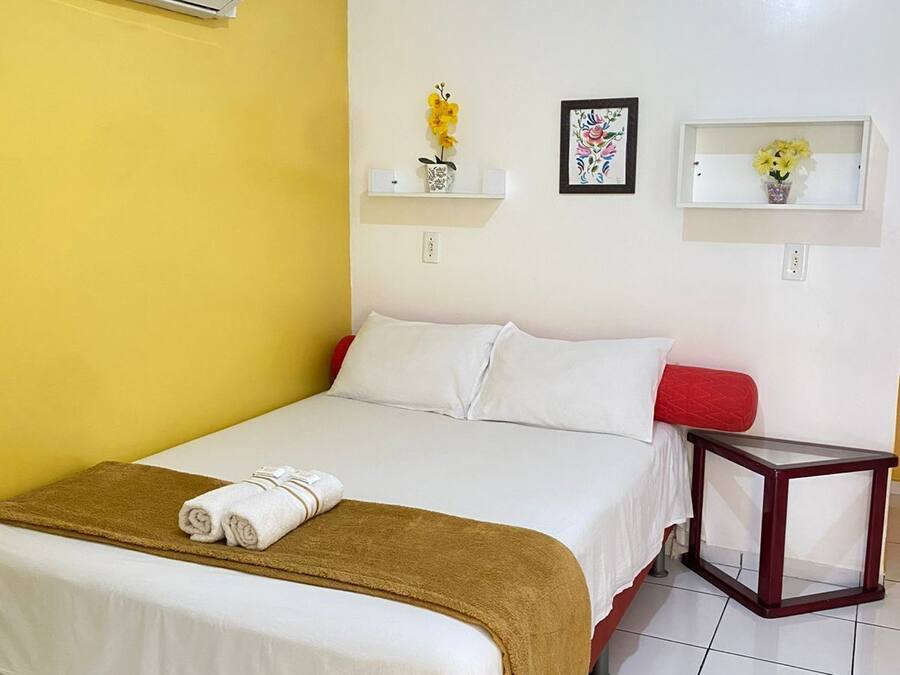 A hotel room with a bed, a bedside table, a wall-mounted air conditioner, and two wall-mounted shelves with decorative items.