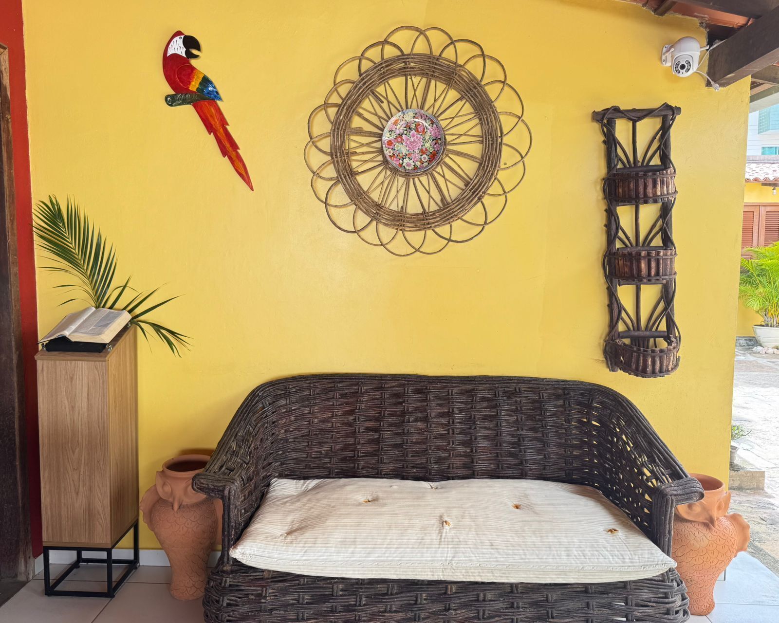 Un sofá de mimbre con un cojín, un adorno circular en la pared, una cámara de seguridad instalada y una decoración de loro.
