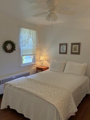 A bedroom with a bed, a nightstand, a lamp, a window with lace curtains, a ceiling fan, and two framed pictures on the wall. - Charming Seashore Cottage • Gardens Location • Beach Tags Included (Ocean City)