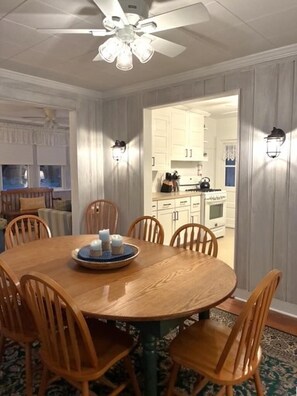 A dining room with a wooden table and chairs, a ceiling fan, and a view into the kitchen. - Charming Seashore Cottage • Gardens Location • Beach Tags Included (Ocean City)