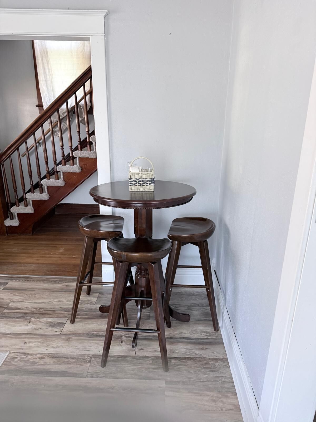 A dining area with a round table and four stools, a wicker basket on the table, and a staircase in the background.