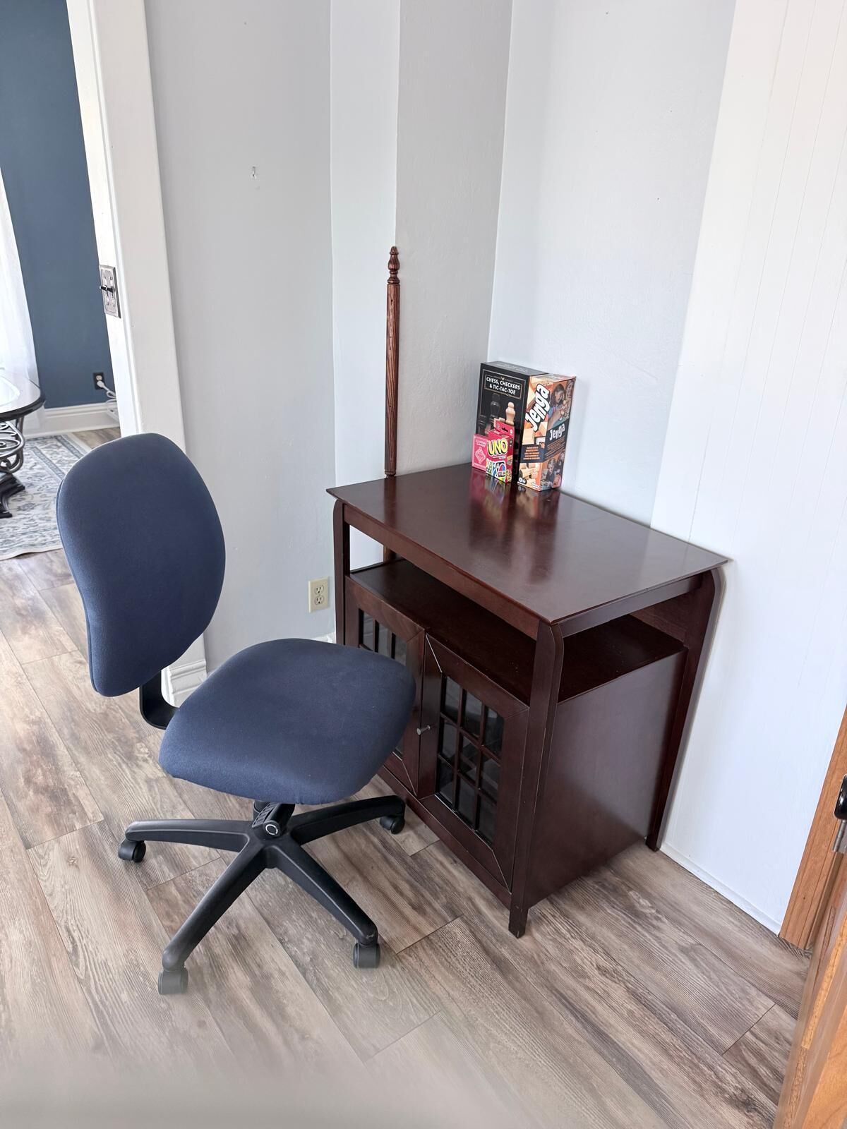 A corner office with a dark wooden desk, a blue office chair with a five-point base, and two board games on the desk.