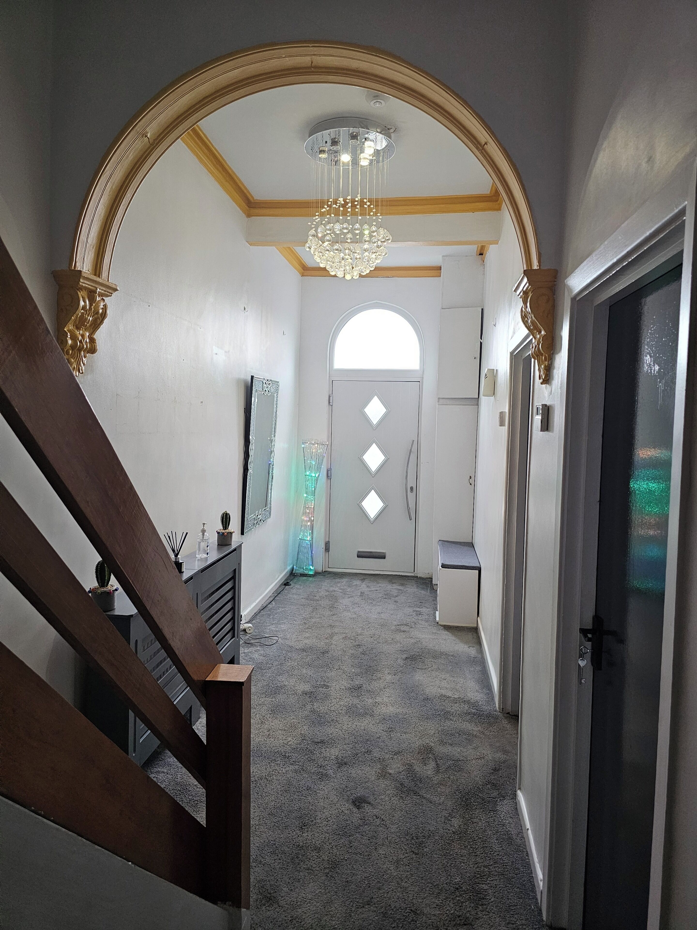 Ein Korridor mit einem imposanten Bogen, einer Chandelüster und einer Tür mit diamantförmigen Fenstern sowie einer Treppe.