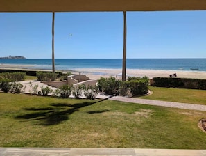 Blick auf einen Strand von einer überdachten Terrasse aus, mit einem Pfad zum Wasser, Palmen und einer Flagge am Boden.