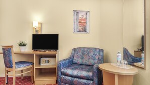 A hotel room with a TV, a patterned armchair, a small table with a glass and water bottle, a chair, and a wall-mounted picture. - XZLLENZ light, parking, 24h digital check in and access, pet friendly (Oberasbach)