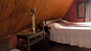 A bedroom with a sloped ceiling, a bed with a white bedspread, a wooden table with a lamp, and a mirror on the wall. - Le Karthala (Antananarivo)
