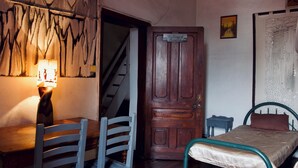 A room with a wooden door, a bed with a green metal frame, a chair, a small table, and a wall with a painting. - Le Karthala (Antananarivo)