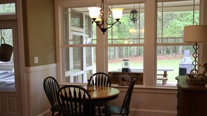 A dining area with a round table, four chairs, a chandelier, and large windows with a view of the outdoors.