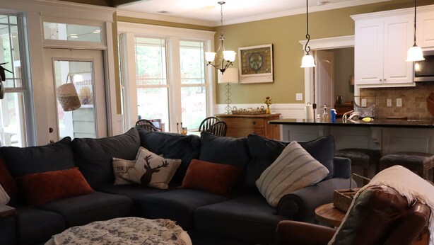 A living room with a sectional sofa, a round ottoman, a wooden coffee table, and a dining area with a kitchen in the background.