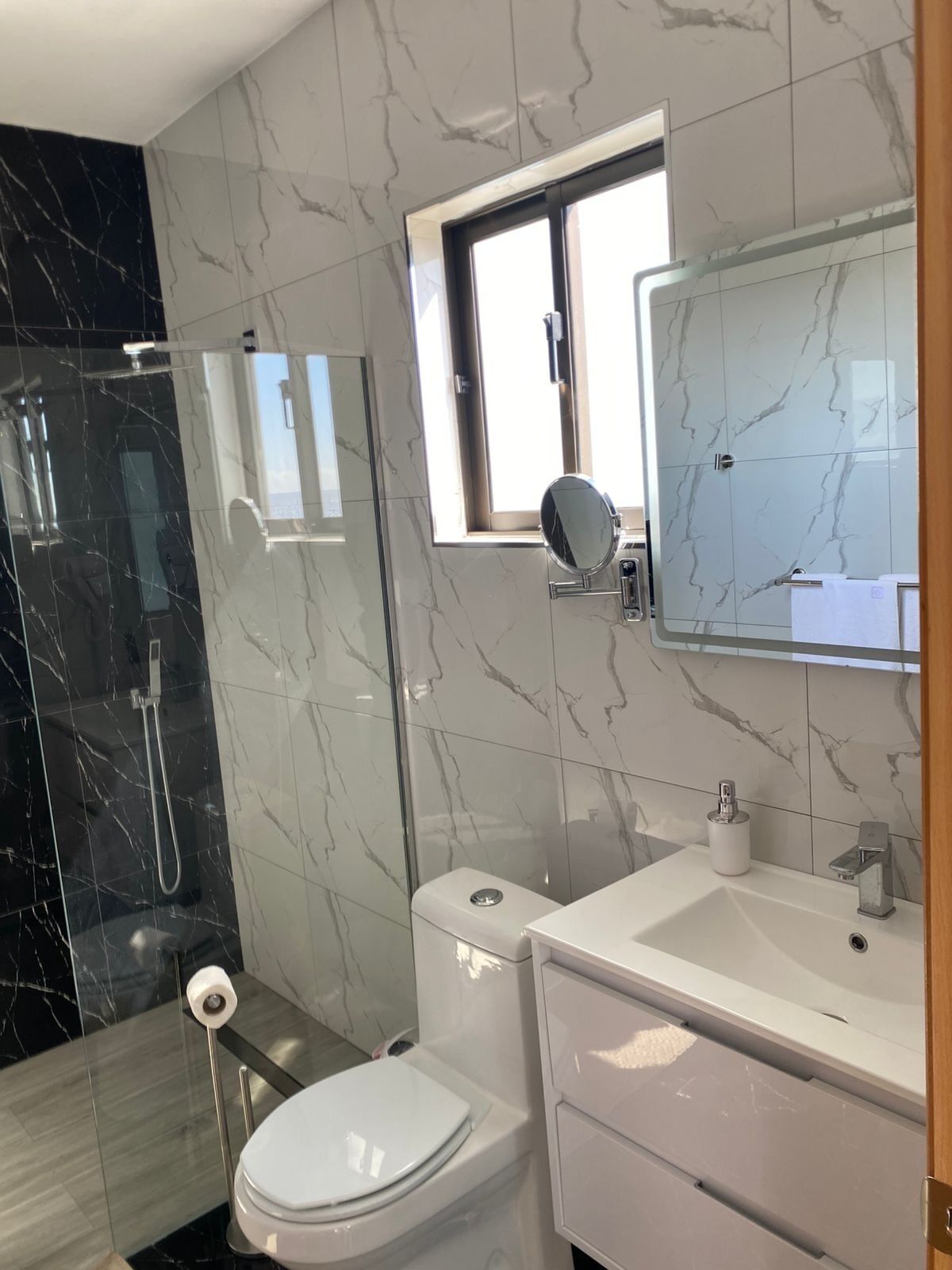 Un baño moderno con cabina de ducha de vidrio, inodoro, lavamanos con espejo y ventana sobre el espejo.