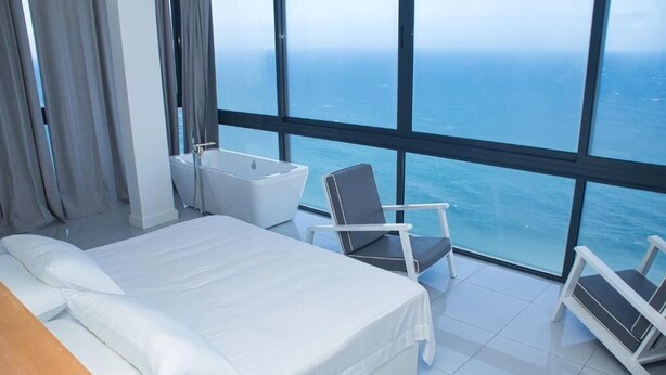 A modern hotel room with a large bed, a bathtub, and a chair by a large window overlooking the sea. - The View Hotel CV (Sao Filipe)