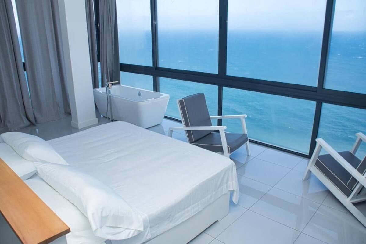 A modern hotel room with a large bed, a bathtub, and a chair by a large window overlooking the sea.
