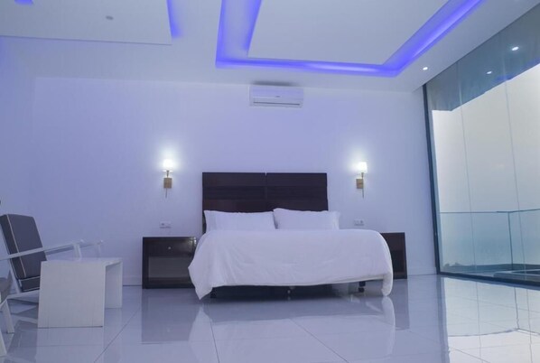 A modern hotel room with a large bed, bedside lamps, a chair, a small table, and a glass wall.