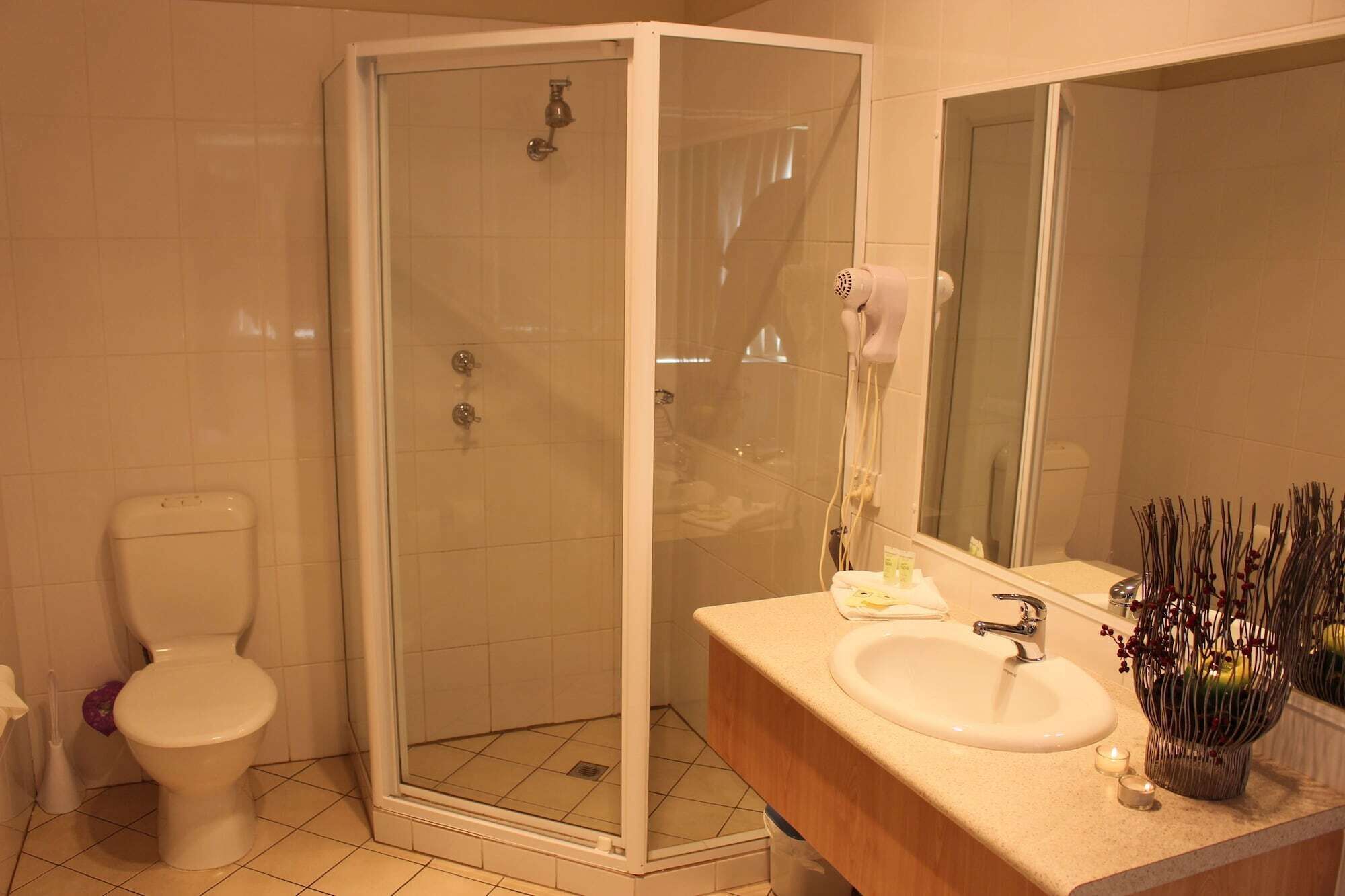 A bathroom with a glass-enclosed shower, a toilet, a sink with a mirror, and a vase with dried flowers.