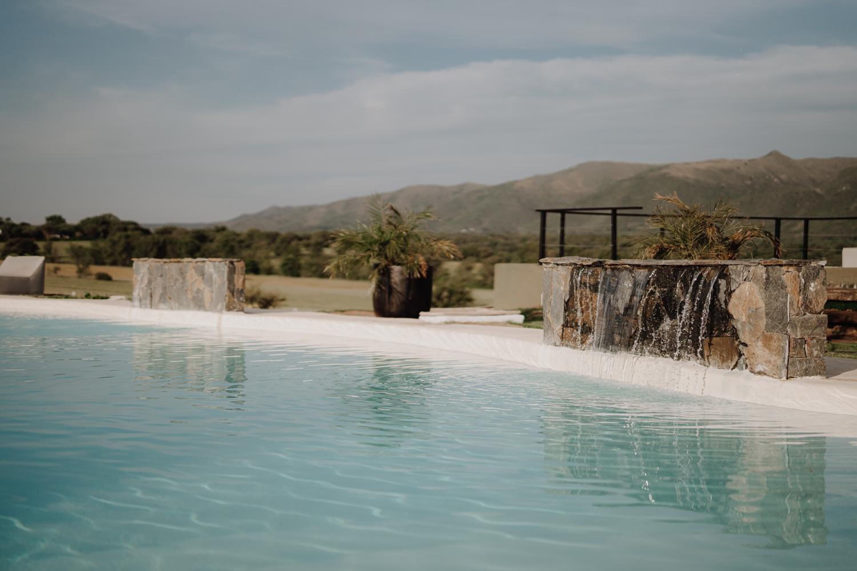 Una piscina con cascada, rodeada de estructuras de piedra y palmeras, con montañas al fondo.