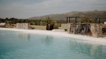 Una piscina con cascada, rodeada de estructuras de piedra y palmeras, con montañas al fondo.