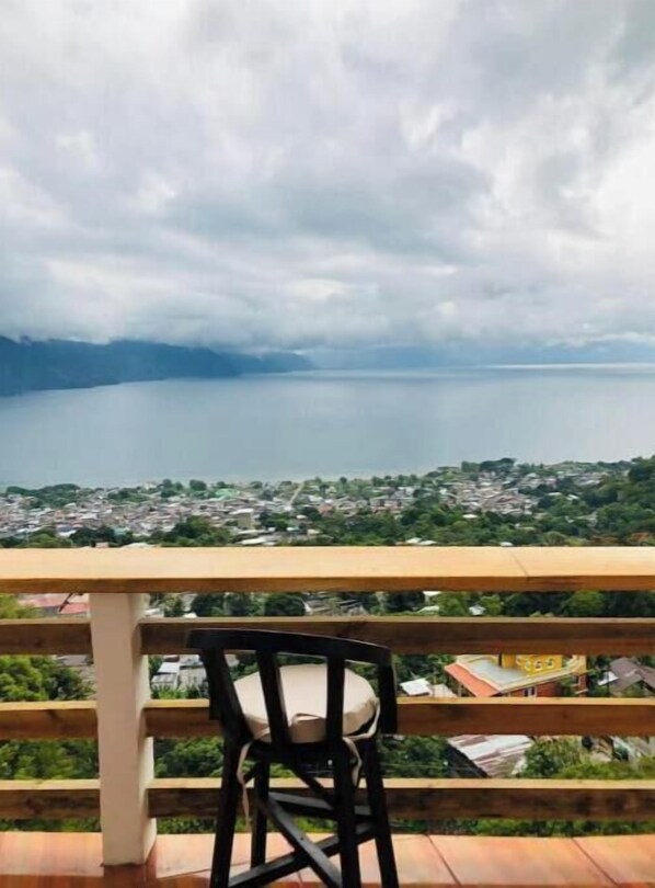 A balcony with a wooden railing, a single black chair, and a view of a lake and a cityscape. - Sunrise High Over Lake 2 (San Pedro La Laguna)