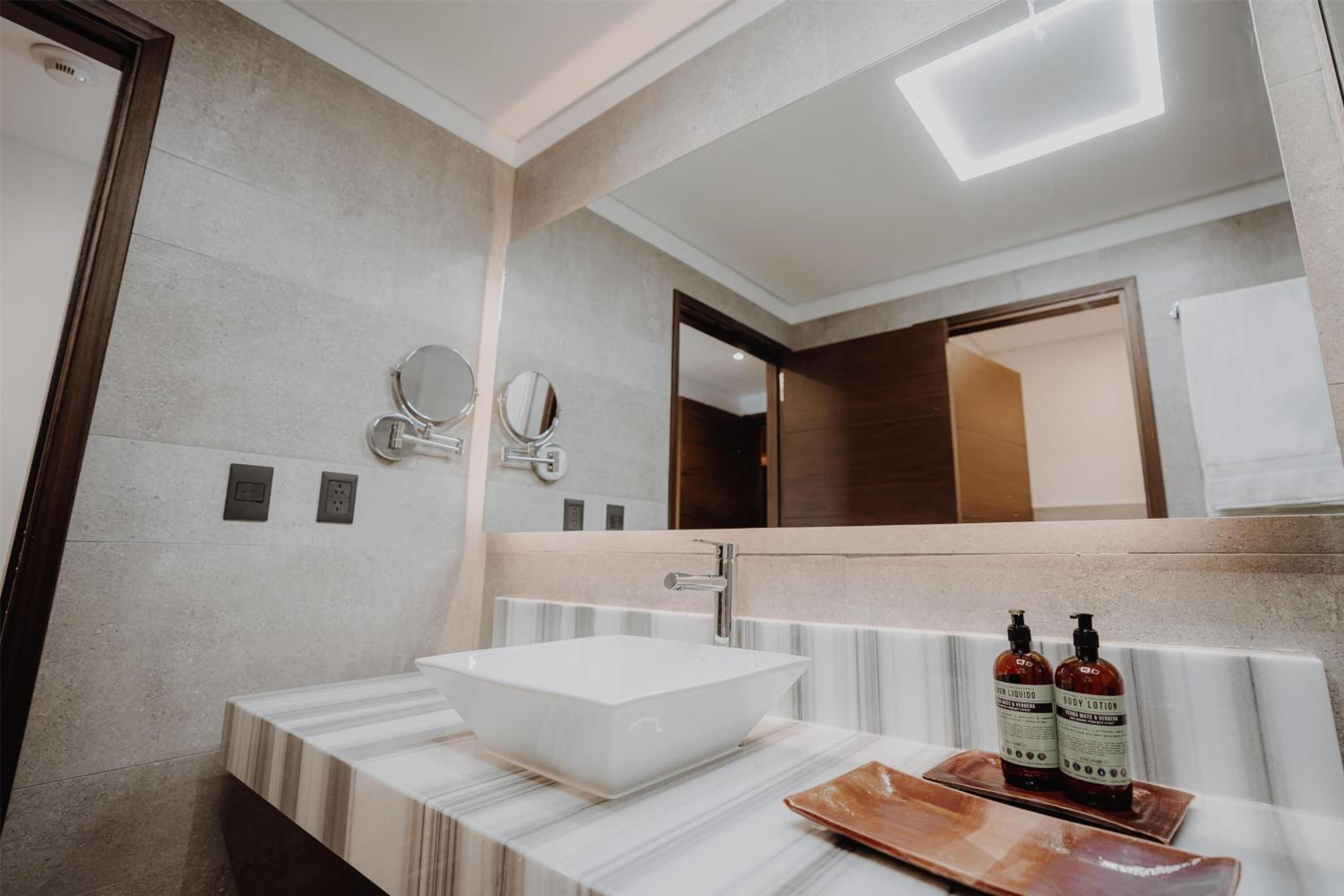 Ein modernes Badezimmer mit einem großen, rechteckigen Waschbecken, zwei wandmontierten Spiegeln und einem rechteckigen Spiegel über dem Waschbecken. Auf der Ablagefläche stehen zwei Flaschen Körperlotion.