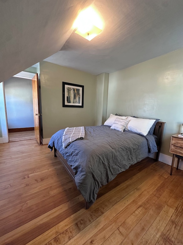 A bedroom with a bed, wooden floor, a ceiling light, a framed picture, and a door leading to another room.
