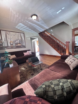 A spacious living room with a large sofa, a coffee table, and a laptop on a stand. A staircase leads to another level, and a painting is on the wall.