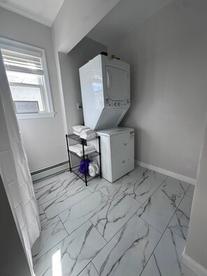 A laundry room with a stacked washer and dryer, a shelf with folded towels, and a window with blinds.