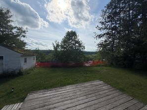 A wooden deck, a white building, and a fenced area with trees in the background. - Family-Friendly 3BR Home Burke Mountain & Kingdom Trails Quiet, Sunlit, Sleeps 6 (Lyndon)