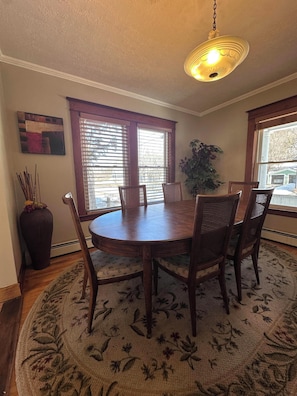 A dining area with a round table, six chairs, a hanging light fixture, a large window with blinds, a potted plant, and a decorative vase. - Family-Friendly 3BR Home Burke Mountain & Kingdom Trails Quiet, Sunlit, Sleeps 6 (Lyndon)