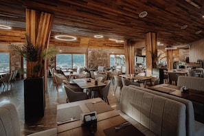 Un restaurant moderne avec des panneaux de plafond en bois, de grandes fenêtres offrant une vue dégagée et différents types de sièges, notamment des tables et des banquettes.