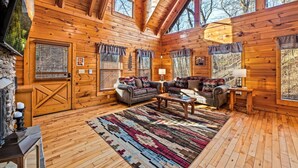 L’intérieur d’une cabane en rondins, avec une cheminée, un plancher en bois, un grand tapis et un ensemble de canapés.