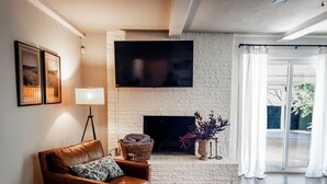 En stue med en brun lædersofa, et fladskærms-tv monteret på en hvid murstensvæg, en pejs og udsigt gennem store glasdøre.
