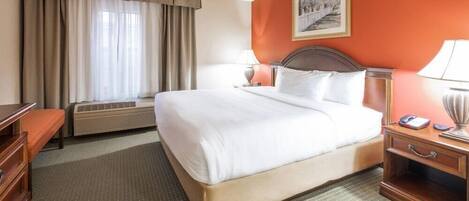 A hotel room with a bed, bedside tables, a dresser, a window with curtains, an air conditioning unit, and a framed picture on the wall.