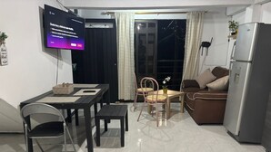 Ein Wohnzimmer mit Fernseher, Esstisch mit Stühlen, einer Couch, einem Kühlschrank und einer Glastür zum Balkon.