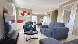 A living room with a sofa, a coffee table, a TV, and a dining area in the background. - Family-Friendly 3BR Home Near Calamvale Central (Calamvale)
