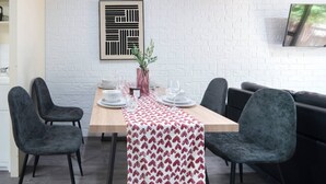 Een eethoek met een witte bakstenen muur, een lijst met abstracte kunst, een gedekte tafel met borden, glazen en een vaas met groene planten.