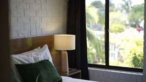 Ein Hotelzimmer mit Bett, Nachttisch mit Lampe, einer Wand mit Backsteinmuster und einem Fenster mit Blick auf Grünflächen.