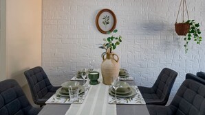 Ein Essbereich mit weißer Ziegelwand, einer hängenden Pflanzkorb mit Grünpflanzen, einer Holzzvase mit einer Pflanze sowie grau gepolsterten Stühlen um einen Tisch, der mit Tellern, Gläsern und einer grünen Schüssel gedeckt ist.