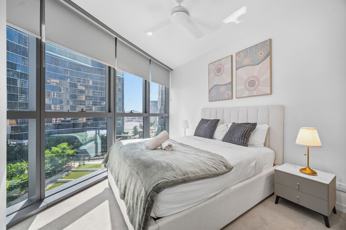 A modern bedroom with a large window, a bed, a nightstand, and two wall art pieces.