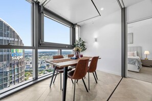 A modern hotel room with a dining area featuring a table and chairs, a large window with a city view, and a separate bedroom with a bed and nightstand. - Luxury 54F Skyline Apt | Iconic Queen’s Wharf 1BR (Brisbane City)