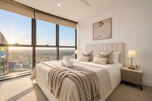 A bedroom with a large bed, two bedside tables, a nightstand, a window with a city view, and a framed artwork on the wall. - Luxury 54F Skyline Apt | Iconic Queen’s Wharf 1BR (Brisbane City)