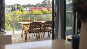 Uma varanda com mesa e cadeiras de madeira, vista para áreas verdes e edifícios, e uma porta de vidro que dá para o interior.