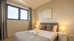 Quarto de hotel com uma cama, duas luminárias de cabeceira, uma janela com cortinas e uma obra de arte na parede.
