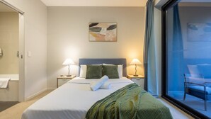 A hotel room with a bed, two bedside lamps, a painting on the wall, and a view through a large window. - Modern 2BR | Ducted A/C, Parking & Great Location (MacGregor)