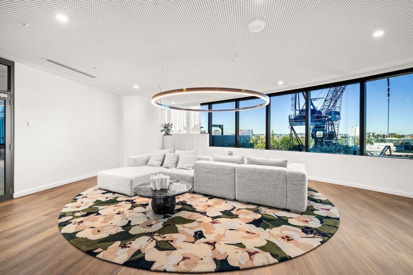 Ein modernes Wohnzimmer mit einem Ecksofa, einem runden Couchtisch und einem großen Fenster mit Blick auf eine Baustelle.